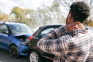 young man rubbing neck in pain from whiplash injury standing by damaged car after traffic accident